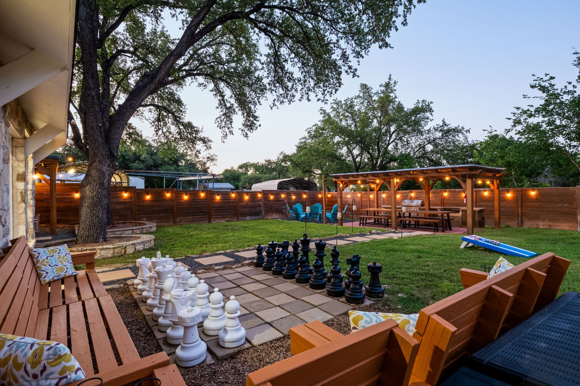 Dragonfly Escape · Backyard with giant chess set, built-in benches, and string lights under oak trees